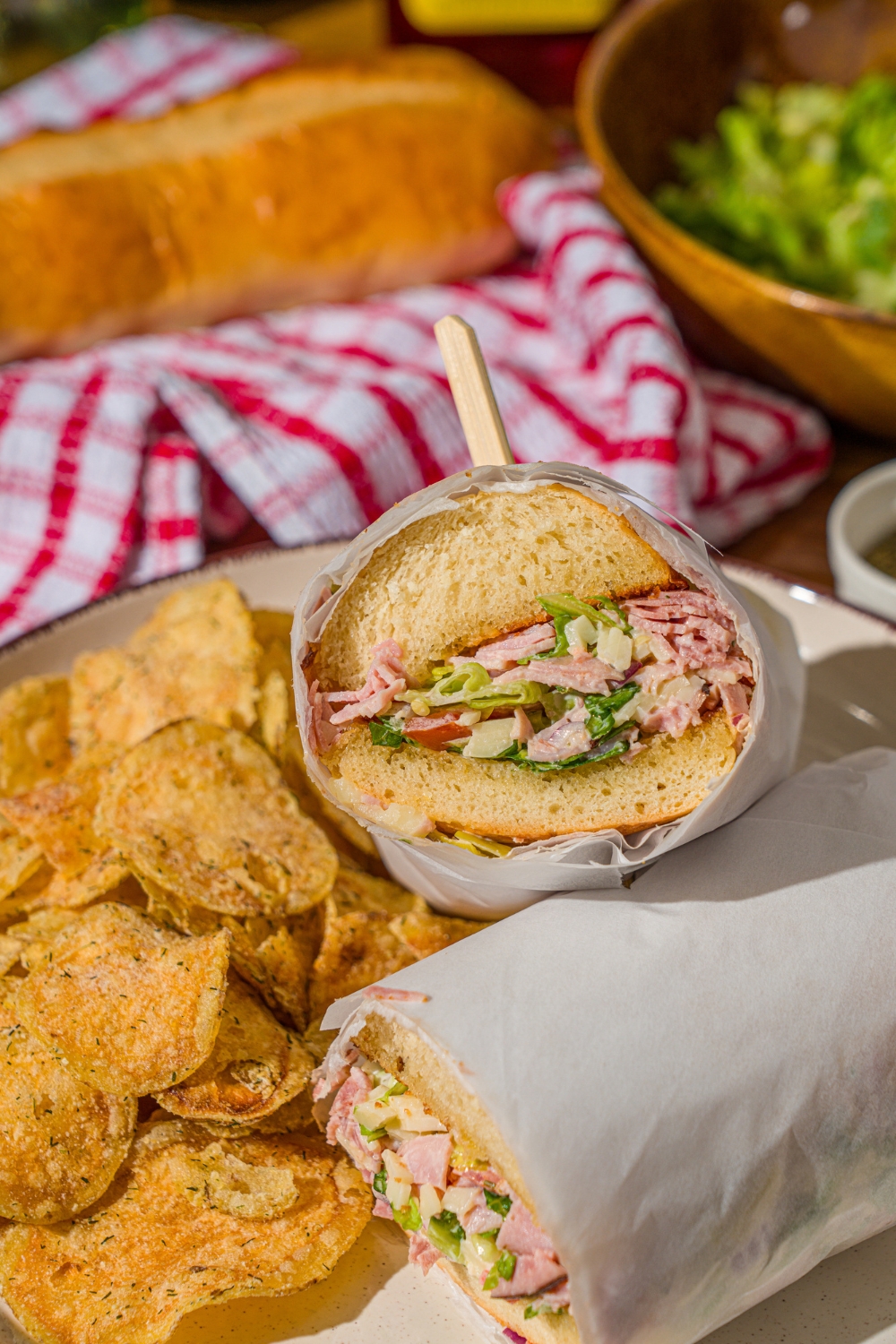 A white plate with a chopped Italian sandwich wrapped in paper and sliced in half, and chips. The plate is on a wooden counter with a bowl of salad and red striped napkin.