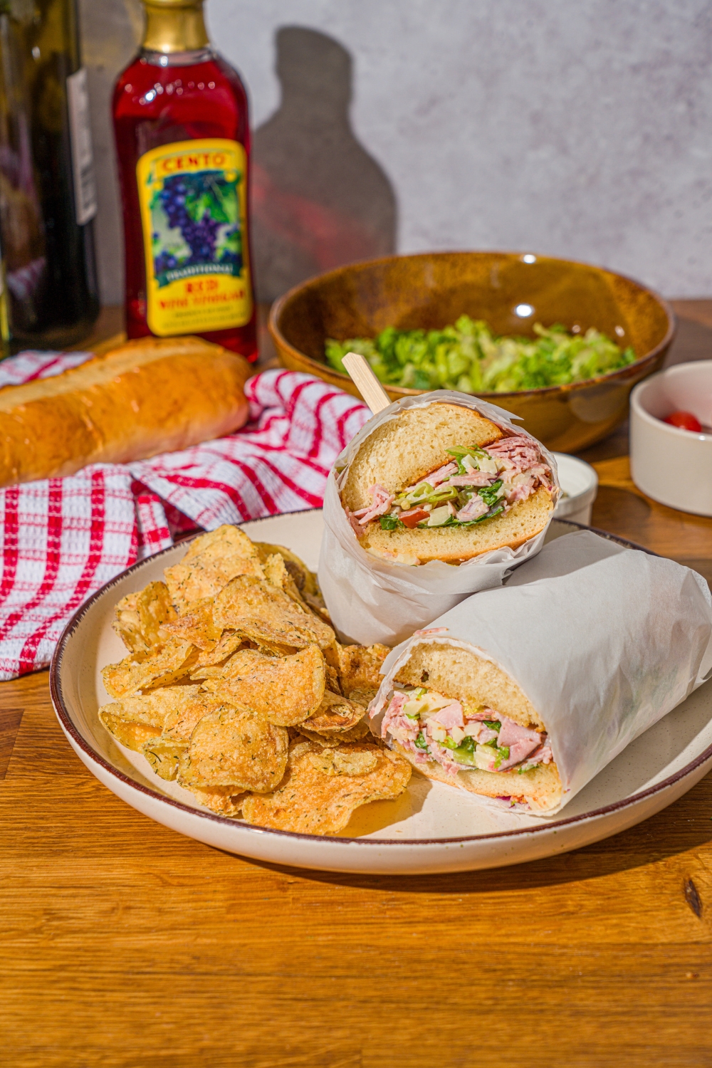 A white plate with a chopped Italian sandwich wrapped in paper and sliced in half, and chips. The plate is on a wooden counter with a bowl of salad and red striped napkin.