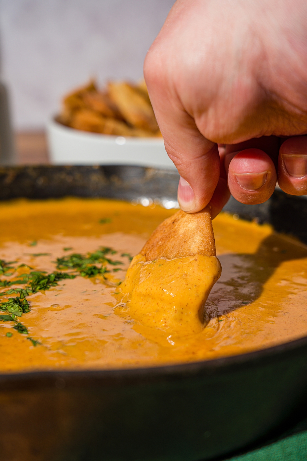 A skillet with Chili's skillet queso garnished with fresh cilantro. A hand is dipping a chip in the queso. The skillet is on a wooden counter with a green cloth napkin and a bowl of chips.