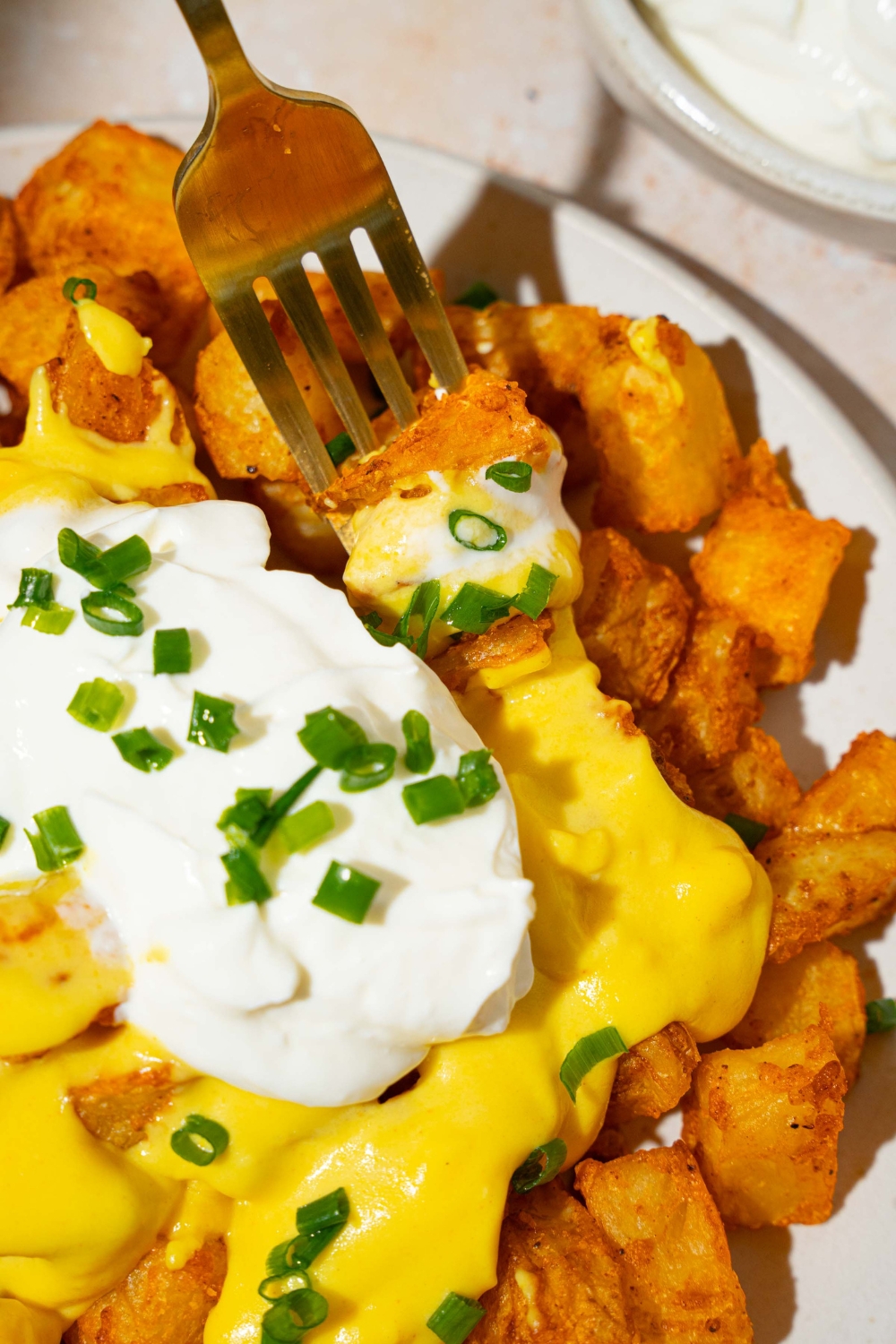 A white plate with copycat cheesy fiesta potatoes topped with taco bell nacho cheese sauce, sour cream, and sliced green onions. There is a fork taking a bite of potatoes. The plate is on a tan counter with a white cloth napkin.