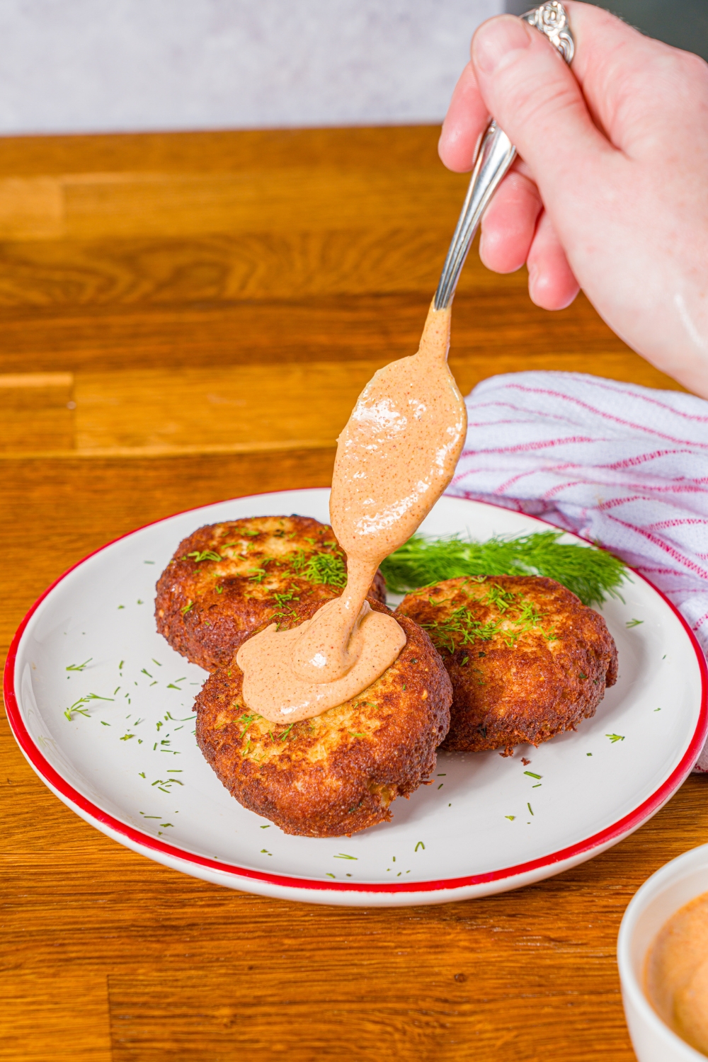 A white plate with crab cakes garnished with fresh dill. A spoon is pouring remoulade sauce on one of the crab cakes. The plate is on a wooden counter.