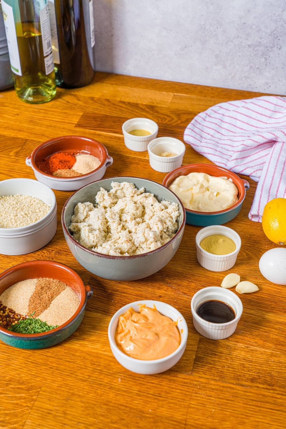A wooden counter with several bowls with ingredients to make crab cakes including lump crab, breadcrumbs, egg, spicy mayo, dijon mustard, Worcestershire sauce, and seasonings.