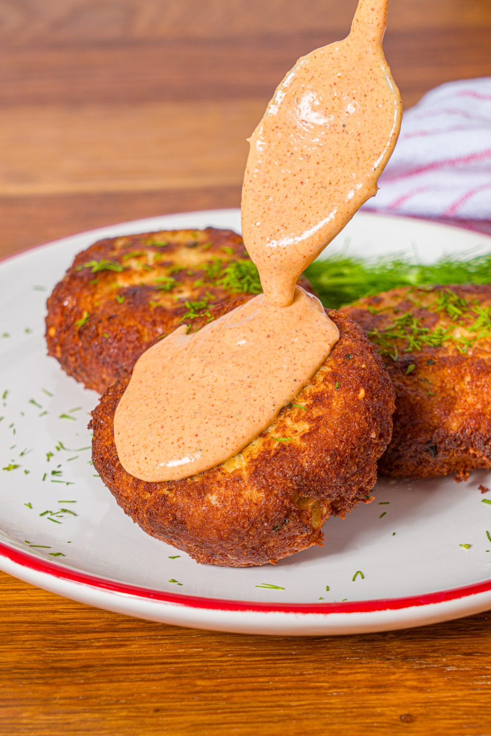 A white plate with crab cakes garnished with fresh dill. A spoon is pouring remoulade on one of the crab cakes. The plate is on a wooden counter.