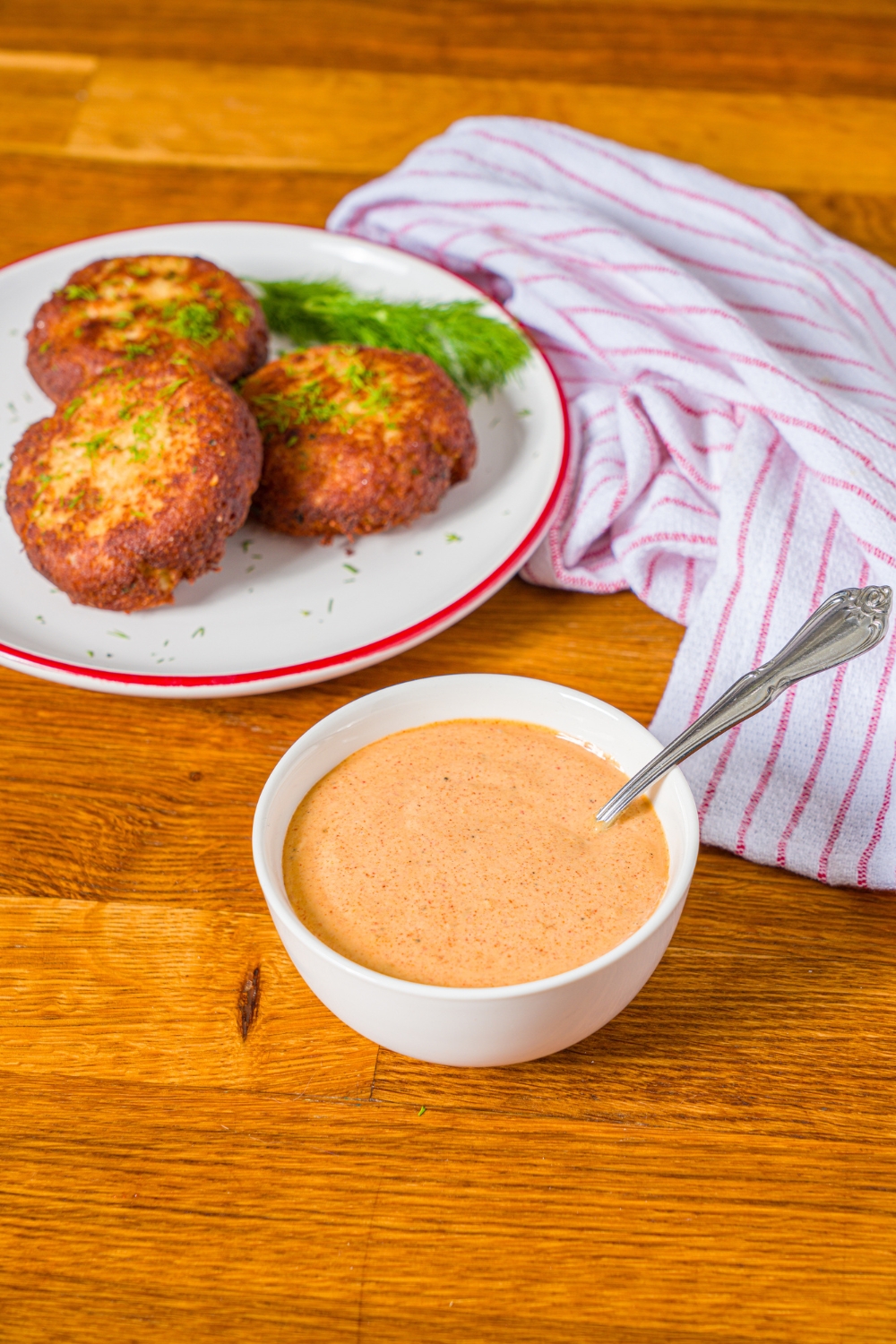 A bowl of crab cake sauce with a spoon in the bowl. The bowl is on a wooden counter with a plate of crab cakes and a red striped napkin.