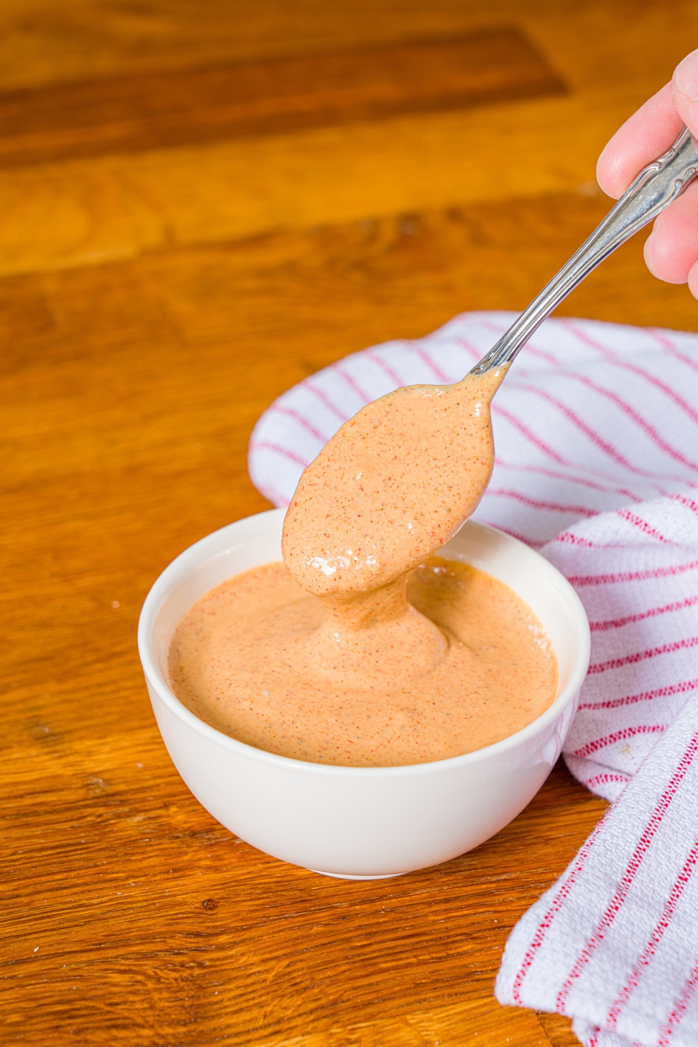 A bowl of crab cake sauce with a spoon dipping into the bowl. The bowl is on a wooden counter with a red striped napkin.