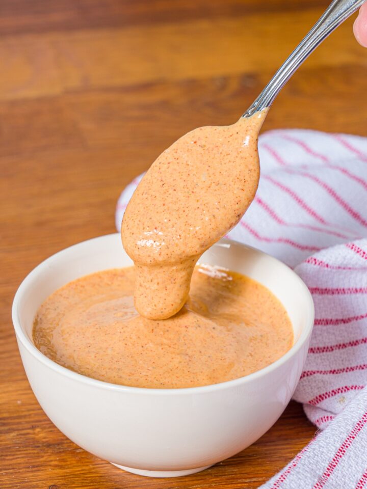 A bowl of crab cake sauce with a spoon dipping into the bowl. The bowl is on a wooden counter with a red striped napkin.