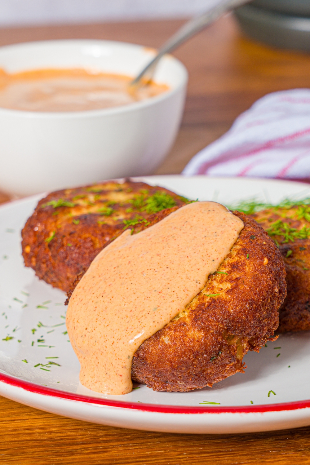 A white plate with crab cakes garnished with fresh dill. One of the cakes is topped with remoulade sauce. The plate is on a wooden counter with a bowl of remoulade sauce.
