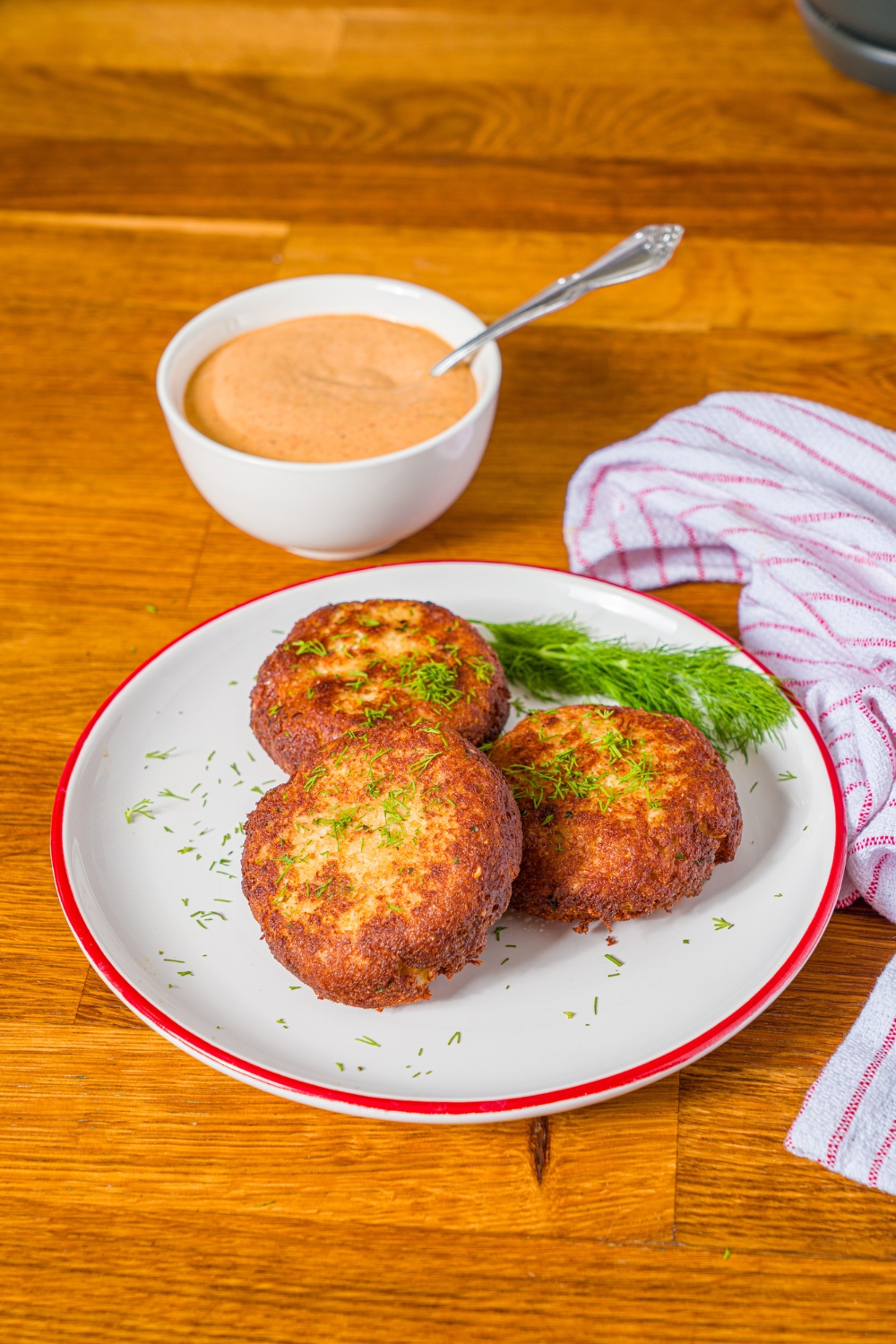 A white plate with crab cakes garnished with fresh dill. The plate is on a wooden counter with a bowl of remoulade sauce.