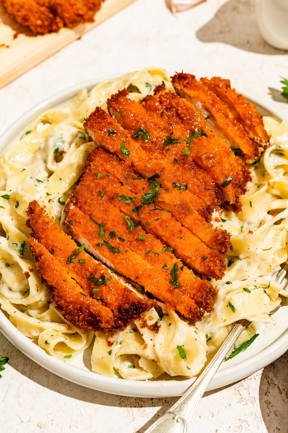 A white plate with crispy Buffalo chicken sliced and served over fettuccine alfredo. The plate is served with a fork and garnished with fresh parsley. The plate is on a tan counter with a board of crispy Buffalo chicken.