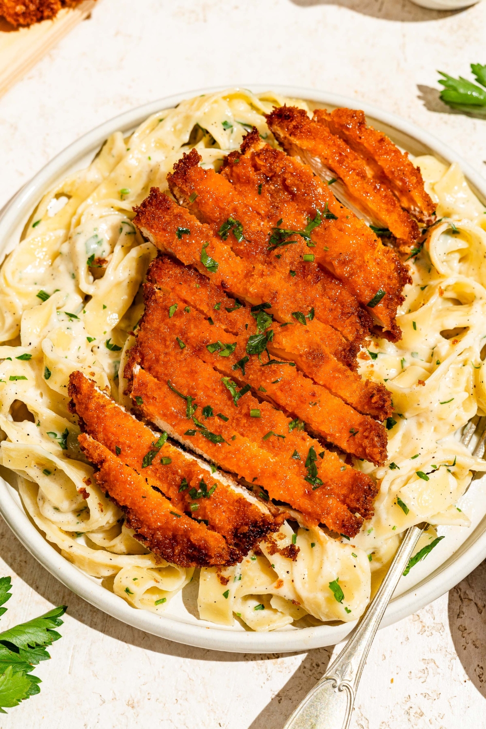 A white plate with crispy Buffalo chicken sliced and served over fettuccine alfredo. The plate is served with a fork and garnished with fresh parsley. The plate is on a tan counter with a board of crispy Buffalo chicken.