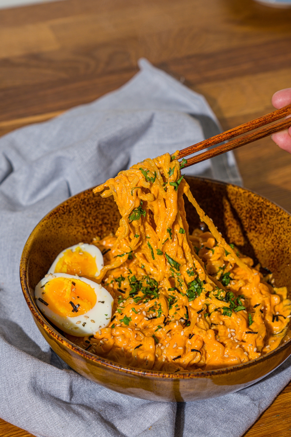 A bowl of Buldak ramen with soft boiled egg and garnished with fresh parsley and sesame seeds. A pair of chopsticks are pulling noodles from the bowl. The bowl is on a wooden counter with a blue cloth napkin.