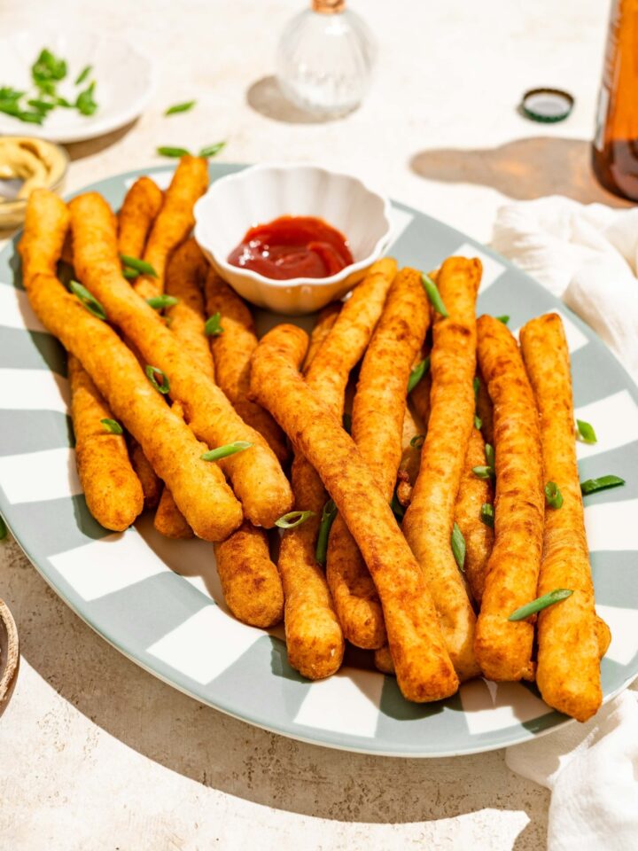 A striped plate with fried Japanese long fries garnished with sliced green onion and served with a small bowl of ketchup. The plate is on a tan counter.