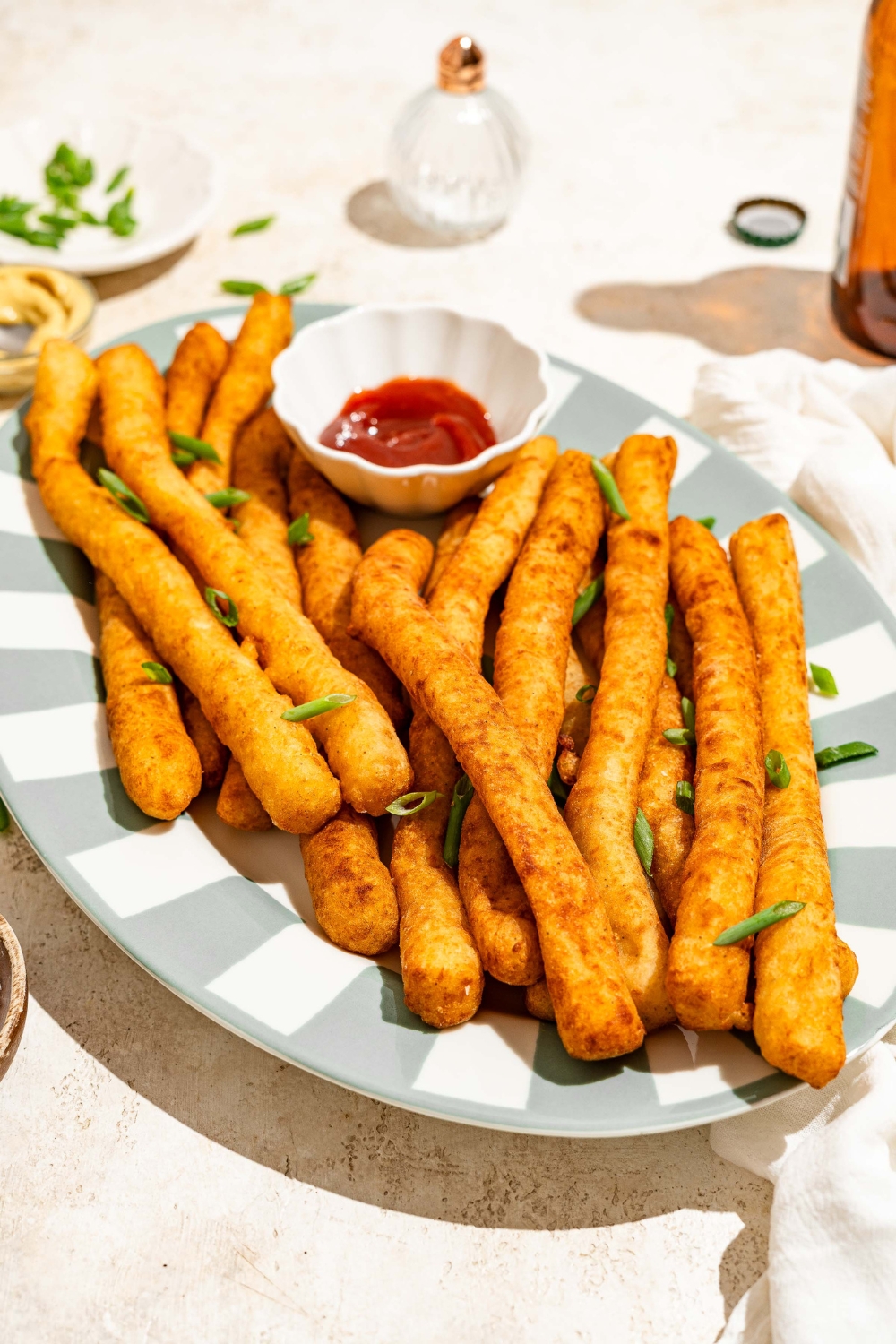 A striped plate with fried Japanese long fries garnished with sliced green onion and served with a small bowl of ketchup. The plate is on a tan counter.