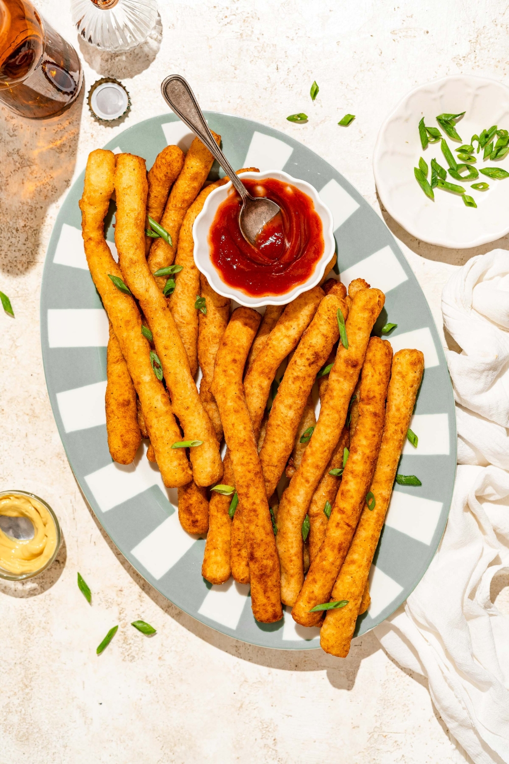 A striped plate with fried Japanese long fries garnished with sliced green onion and served with a small bowl of ketchup. The plate is on a tan counter with small bowl of garnishes.