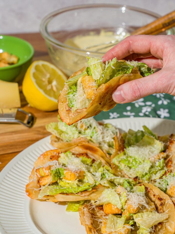 A hand taking a chicken caesar smash taco from a white plate of tacos. The taco is finished with shredded parmesan cheese. The plate is on a wooden counter with a lemon and bowl of caesar dressing.
