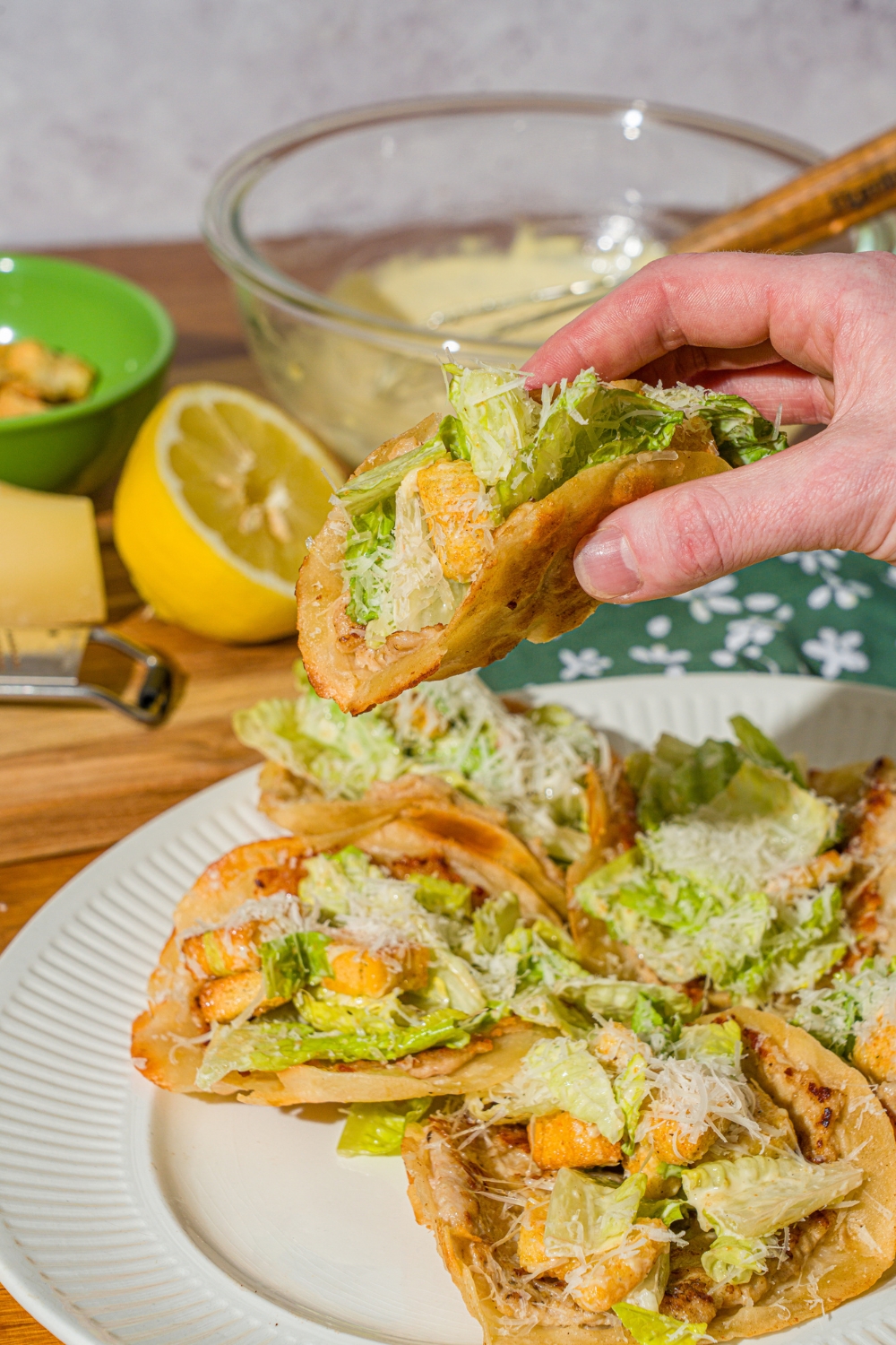 A hand taking a chicken caesar smash taco from a white plate of tacos. The taco is finished with shredded parmesan cheese. The plate is on a wooden counter with a lemon and bowl of caesar dressing.