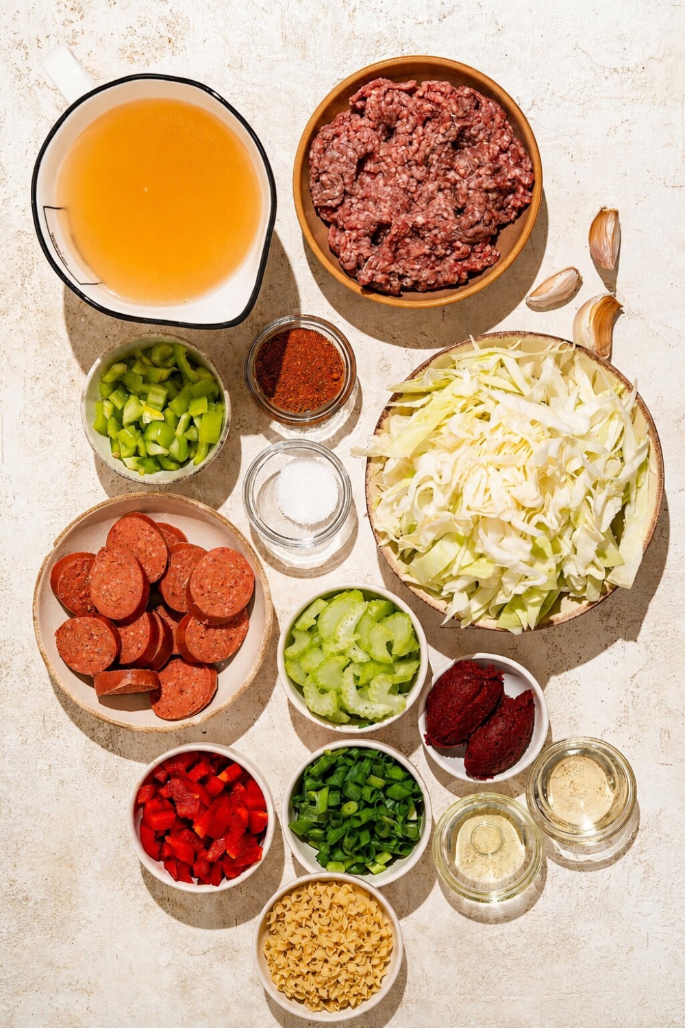 A tan counter with several bowls containing ingredients to make dirty cabbage and noodles including cabbage, ground beef, andouille sausage, chicken stock, celery, bell pepper, scallions, tomato paste, garlic, and more.