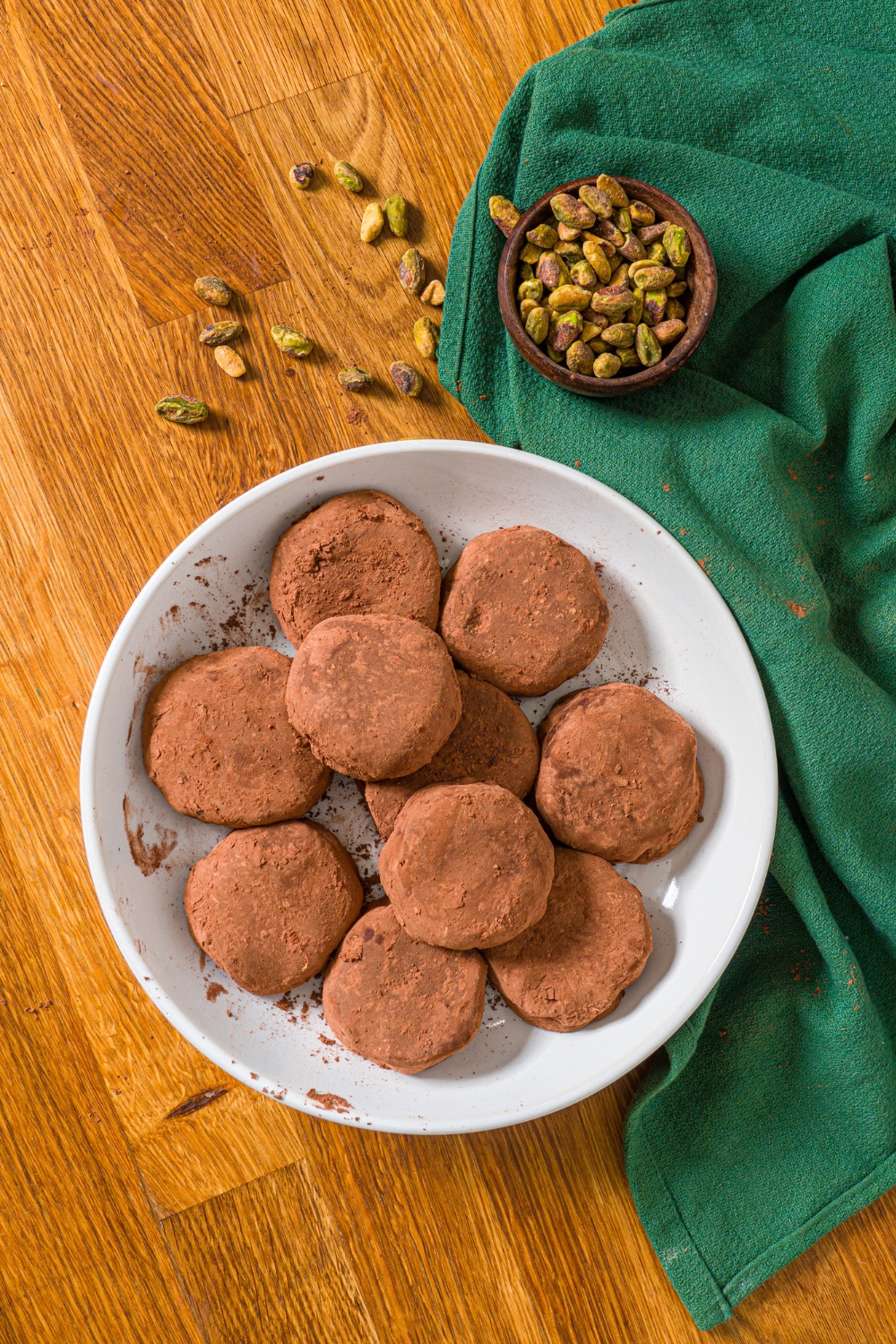 A white plate with Dubai chocolate cookies. The plate is on a wooden counter with a green cloth napkin and a bowl of pistachios.