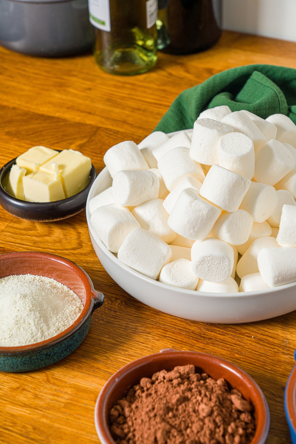 A wooden counter with ingredients to make Dubai chocolate cookies including marshmallows, cocoa powder, butter, and katafi.