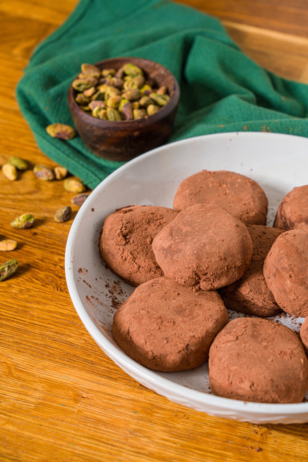 A white plate with Dubai chocolate cookies. The plate is on a wooden counter with a green cloth napkin and a bowl of pistachios.
