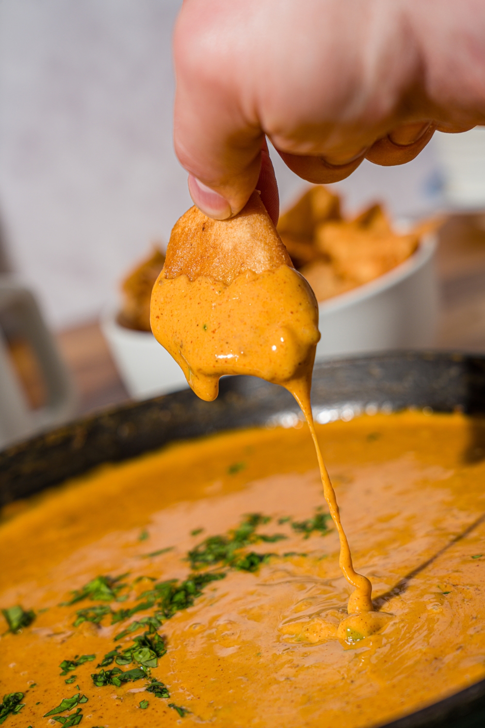 A skillet with Chili's skillet queso garnished with fresh cilantro. A hand is dipping a chip in the queso. The skillet is on a wooden counter with a green cloth napkin and a bowl of chips.