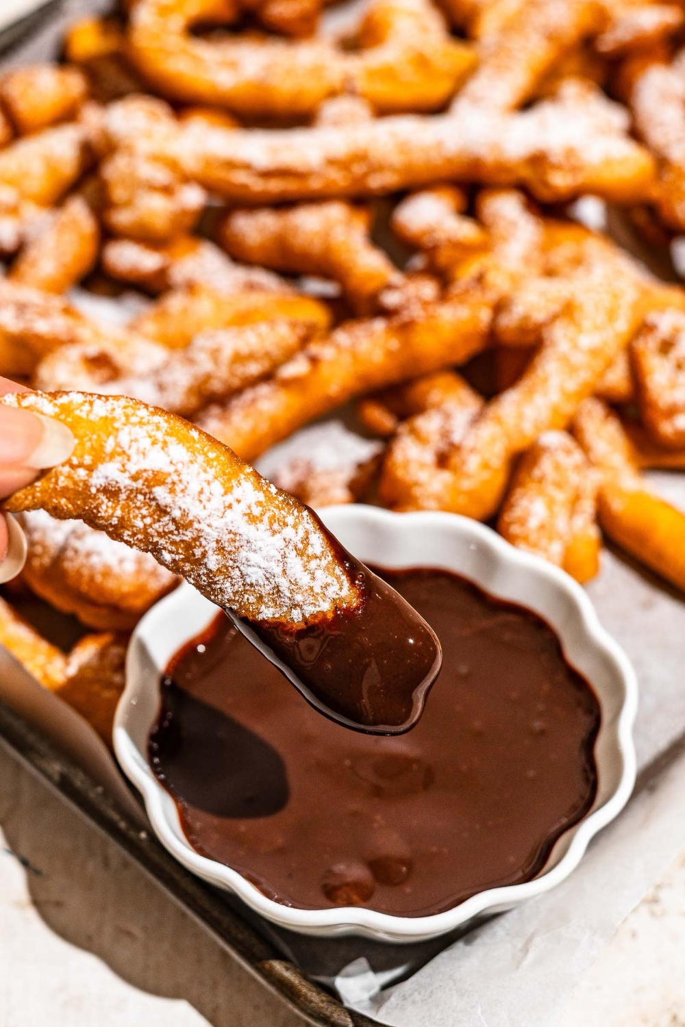 A baking sheet lined with parchment paper with funnel cake fries covered in powdered sugar and served with a bowl of chocolate. A fry is being dipped into the chocolate.