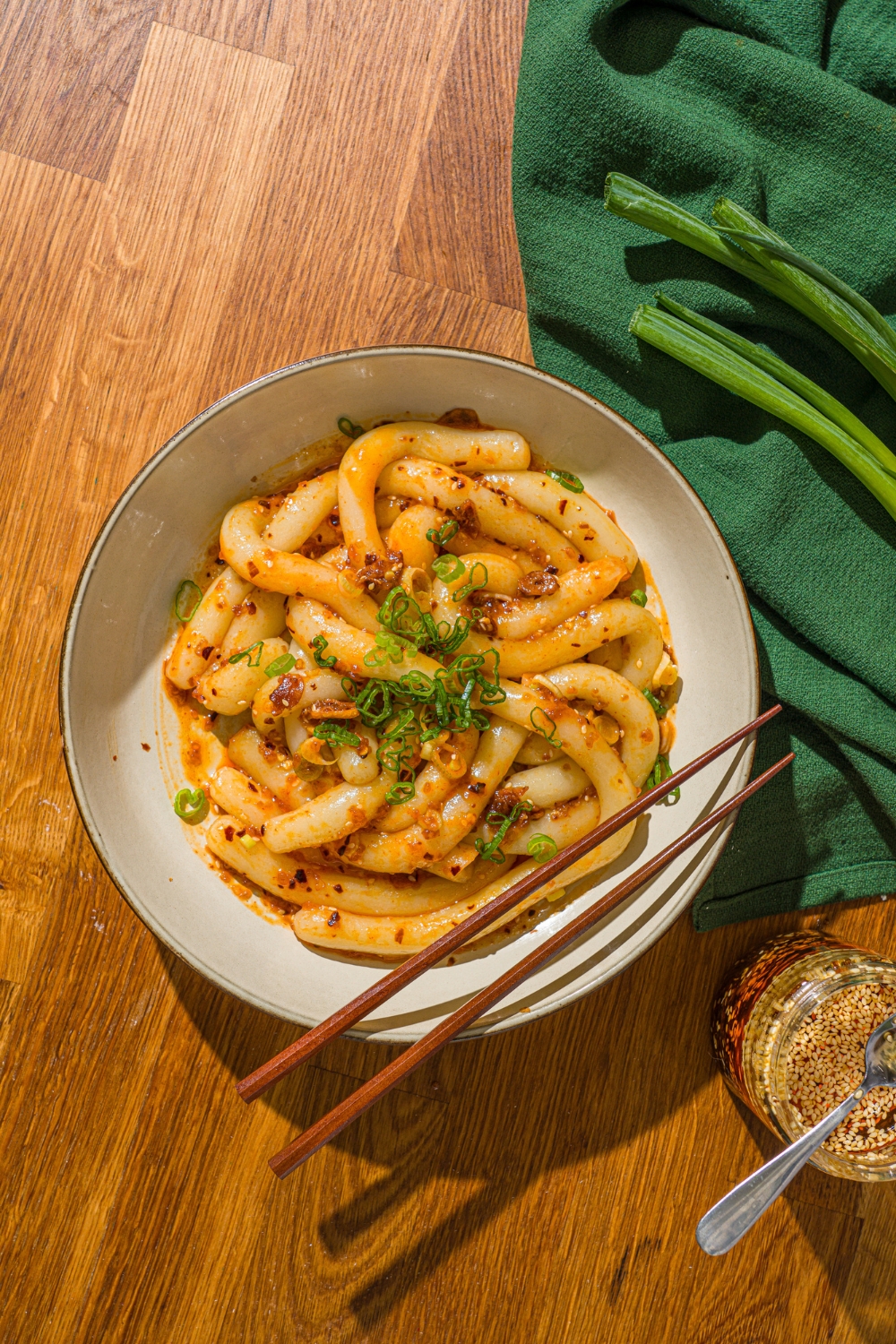 A white bowl with spicy potato noodles tossed in chili crisp and garnished with sliced green onion. A pair of chopsticks are resting on the bowl. The bowl is on a wooden counter with a green cloth napkin.