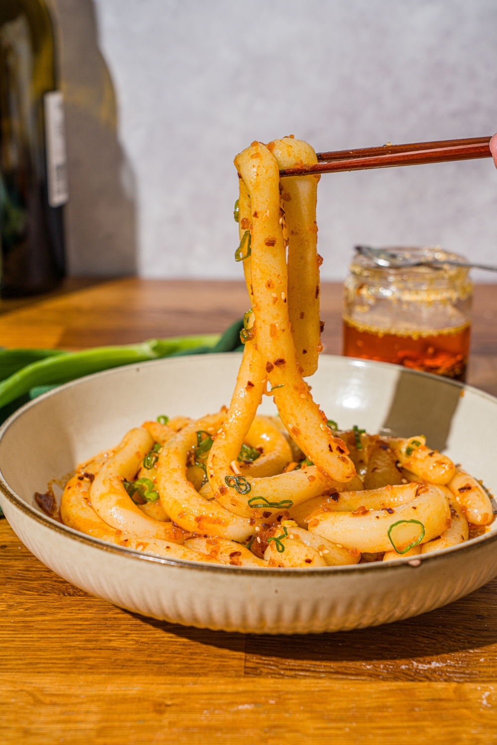 A white bowl with spicy potato noodles tossed in chili crisp and garnished with sliced green onion. A pair of chopsticks are taking a noodle from the bowl. The bowl is on a wooden counter with a jar of chili crisp.