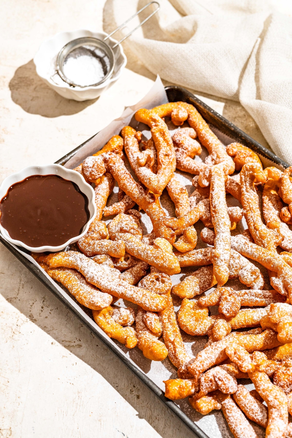 A baking sheet lined with parchment paper with funnel cake fries covered in powdered sugar and served with a bowl of chocolate. The baking sheet is on a tan counter with a bowl of powdered sugar with a sifter.