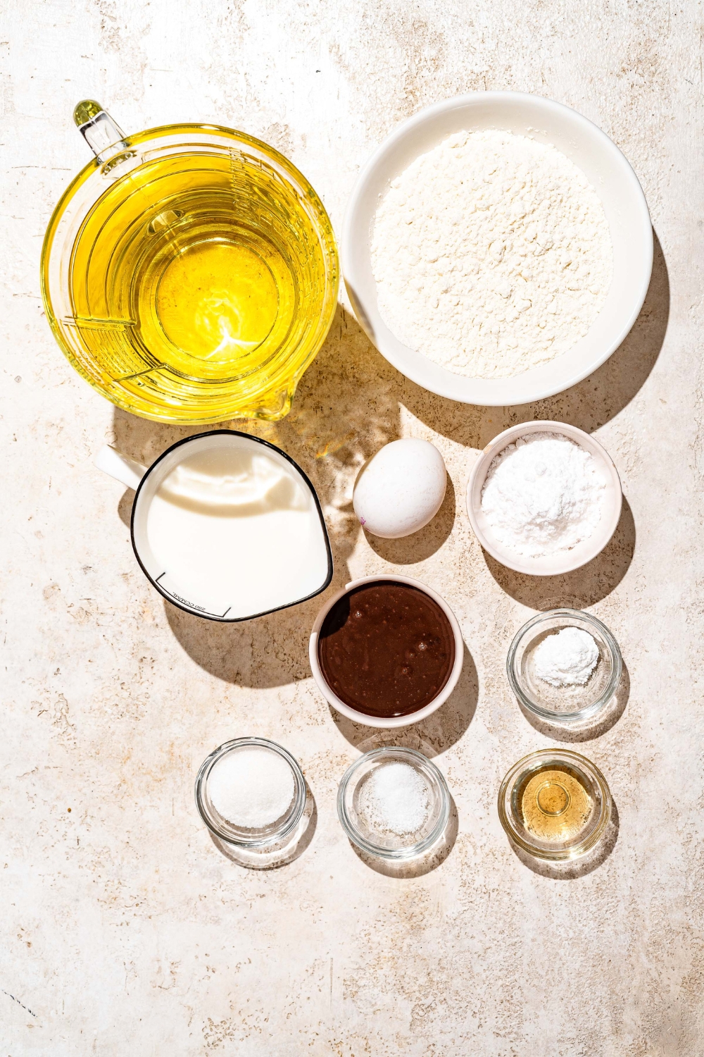 A tan counter with several bowls containing ingredients to make funnel cake fries including oil, flour, sugar, milk, egg, vanilla extract, and powdered sugar.