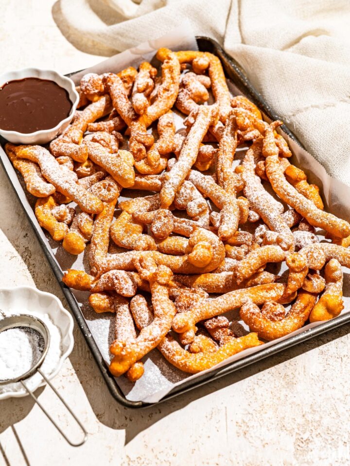 A baking sheet lined with parchment paper with funnel cake fries covered in powdered sugar and served with a bowl of chocolate. The baking sheet is on a tan counter with a bowl of powdered sugar with a sifter.