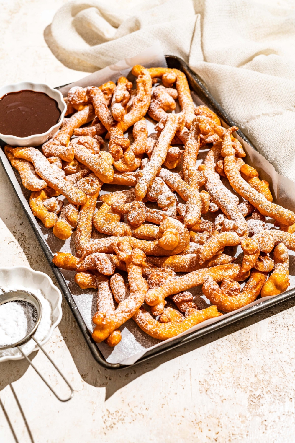 A baking sheet lined with parchment paper with funnel cake fries covered in powdered sugar and served with a bowl of chocolate. The baking sheet is on a tan counter with a bowl of powdered sugar with a sifter.