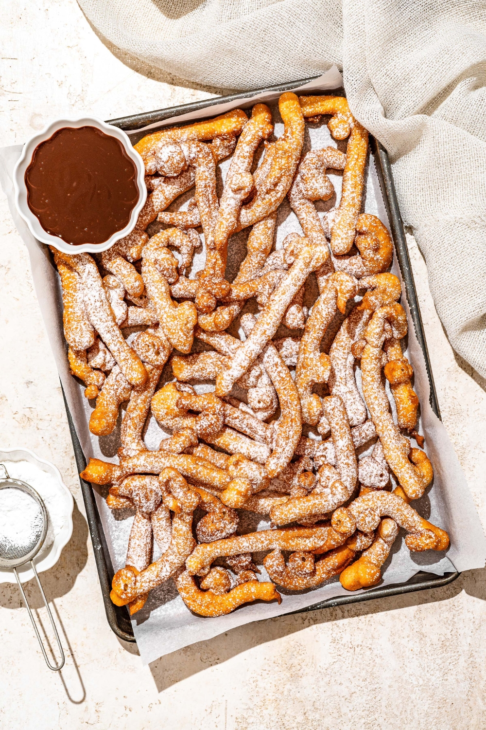 A baking sheet lined with parchment paper with funnel cake fries covered in powdered sugar and served with a bowl of chocolate. The baking sheet is on a tan counter with a bowl of powdered sugar with a sifter.