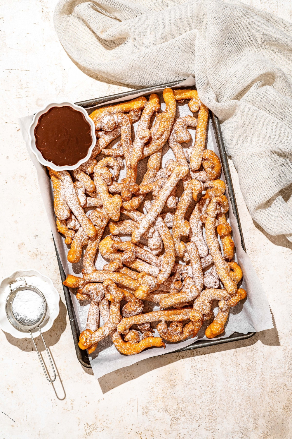 A baking sheet lined with parchment paper with funnel cake fries covered in powdered sugar and served with a bowl of chocolate. The baking sheet is on a tan counter with a bowl of powdered sugar with a sifter.