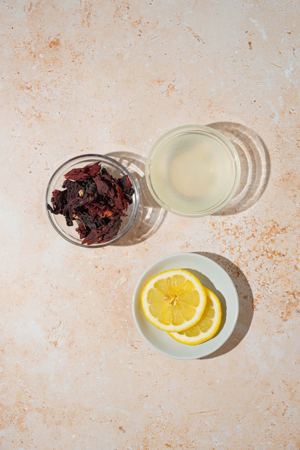 A tan counter with ingredients to make hibiscus lemonade including hibiscus tea leaves, lemonade, and lemon.