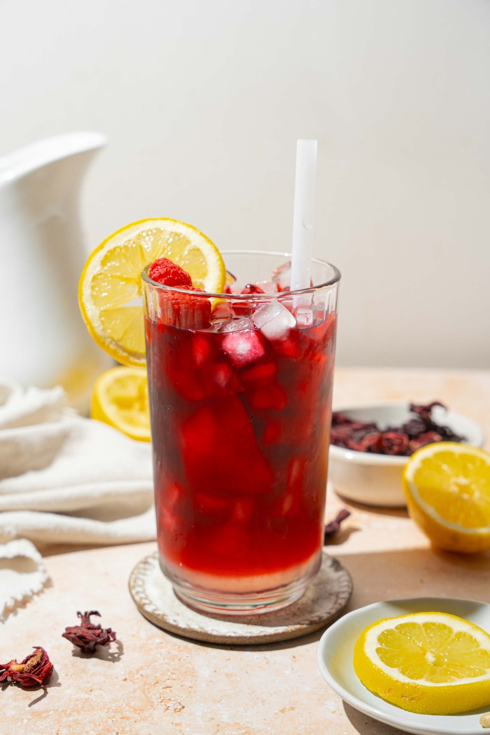 A tall glass of hibiscus lemonade served with ice, sliced lemon, and a straw. The glass is on a coaster on a tan counter with a white cloth napkin, sliced lemon, and a bowl of hibiscus tea leaves.