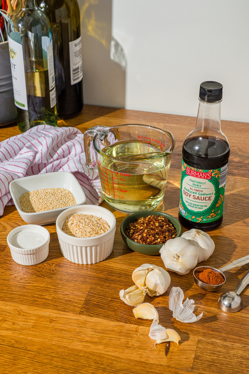 A wooden counter with ingredients to make chili crisp oil including oil, soy sauce, garlic, sesame seeds, minced onion, chili flakes, sugar, salt, and seasonings.