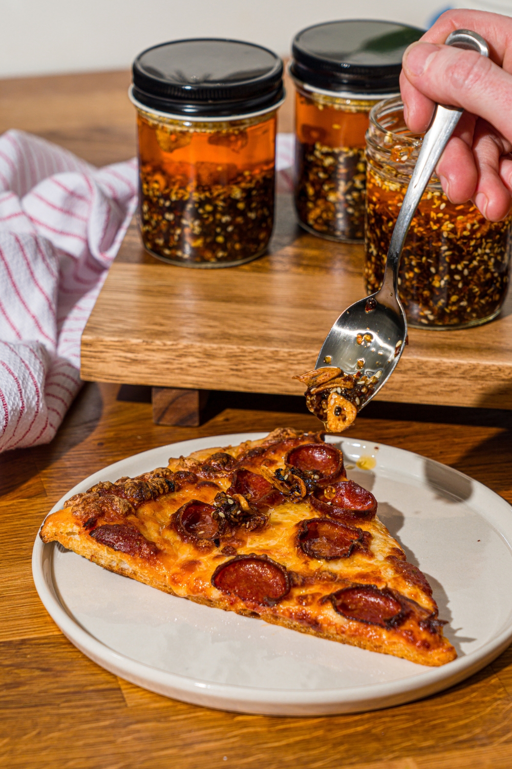 A spoon pouring chili crisp over a white plate with a slice of pepperoni pizza. The plate is on a wooden counter with a board with jars of chili crisp.