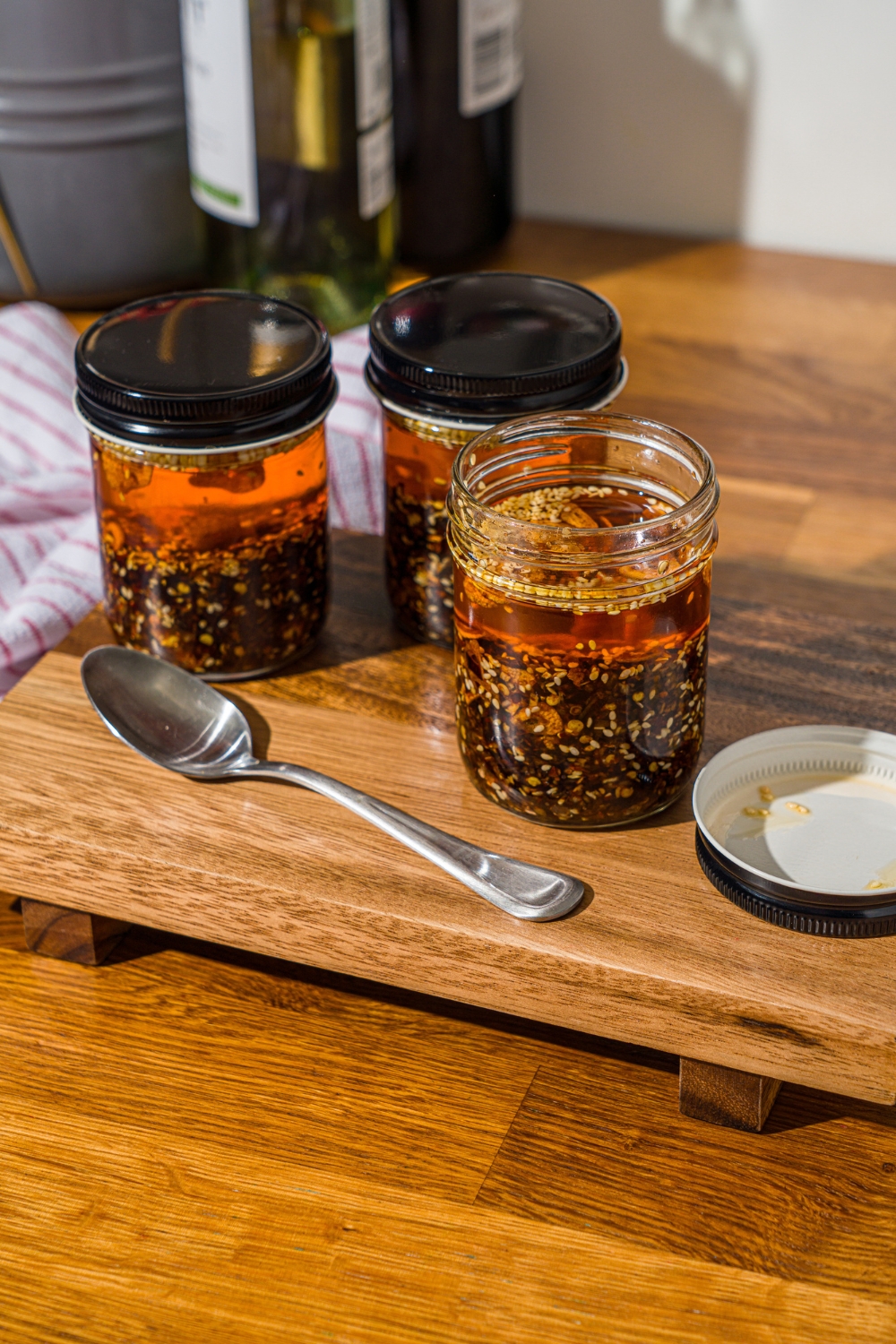 A wooden board with three jars of chili crisp oil. There is a spoon on the board. The board is on a wooden counter.