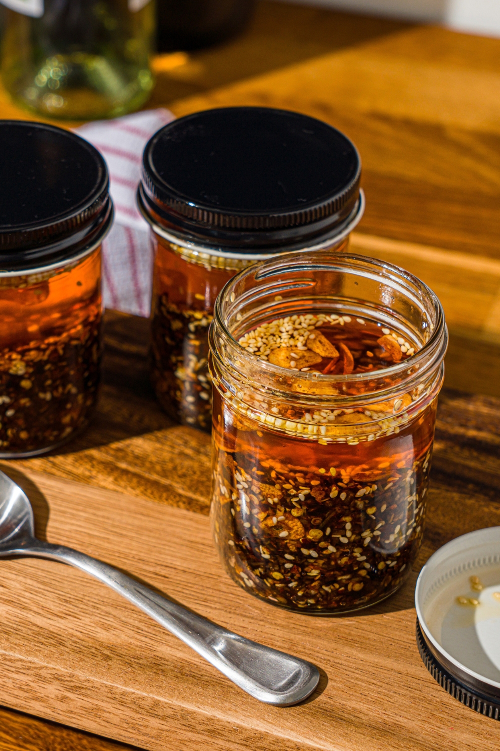 A wooden board with three jars of chili crisp oil. There is a spoon on the board. The board is on a wooden counter.