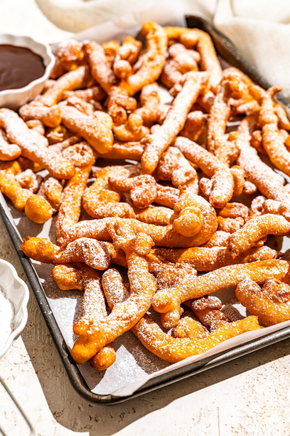 A baking sheet lined with parchment paper with funnel cake fries covered in powdered sugar and served with a bowl of chocolate. The baking sheet is on a tan counter with a bowl of powdered sugar with a sifter.