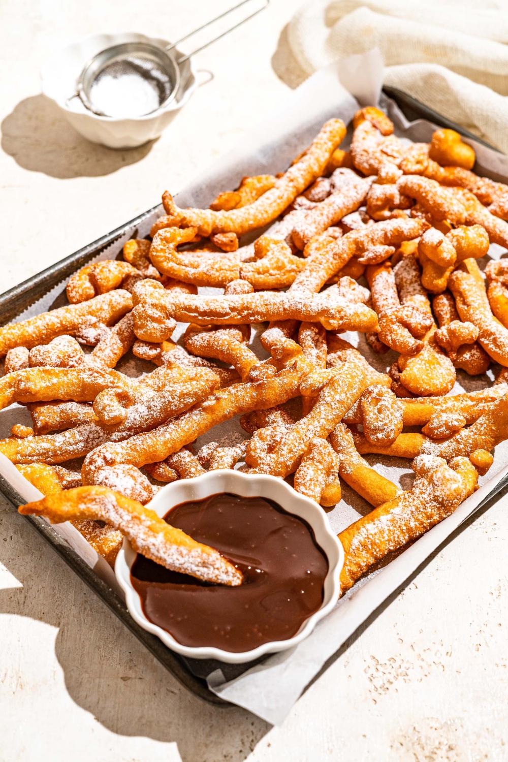 A baking sheet lined with parchment paper with funnel cake fries covered in powdered sugar and served with a bowl of chocolate. A fry is dipped in the chocolate. The baking sheet is on a tan counter with a bowl of powdered sugar with a sifter.