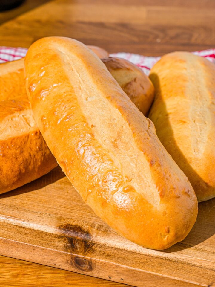 A wooden board with homemade hoagie rolls. The board is on a wooden counter with a red striped napkin.