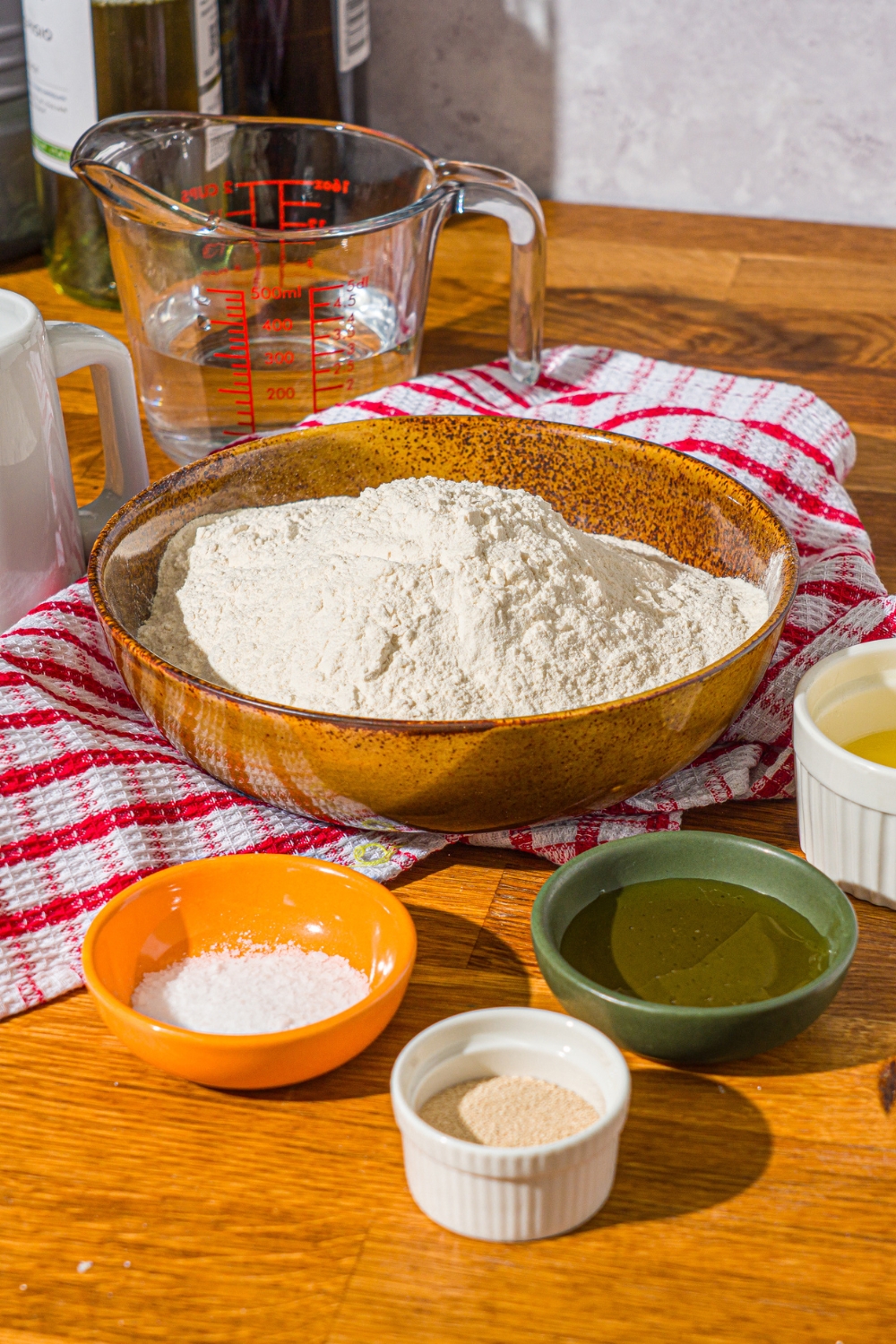 A wooden counter with several bowls containing ingredients to make hoagie rolls including flour, yeast, milk, honey, butter, milk powder, and salt.