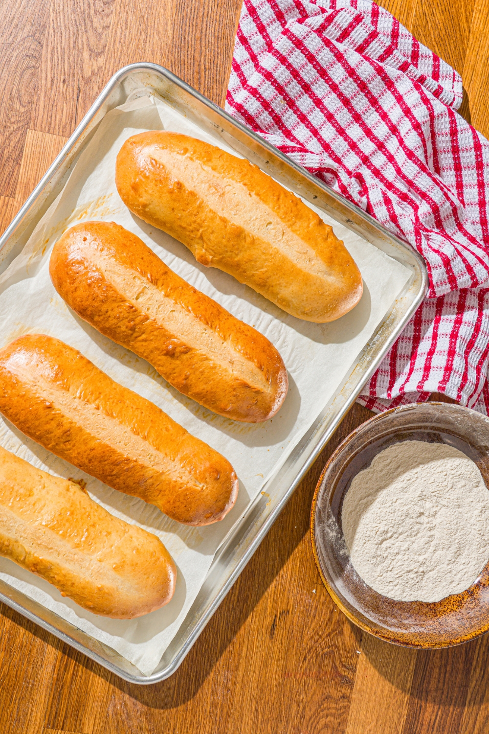 A baking sheet lined with parchment paper with baked homemade hoagie rolls. The sheet is on a wooden counter with a red striped napkin and bowl of flour.