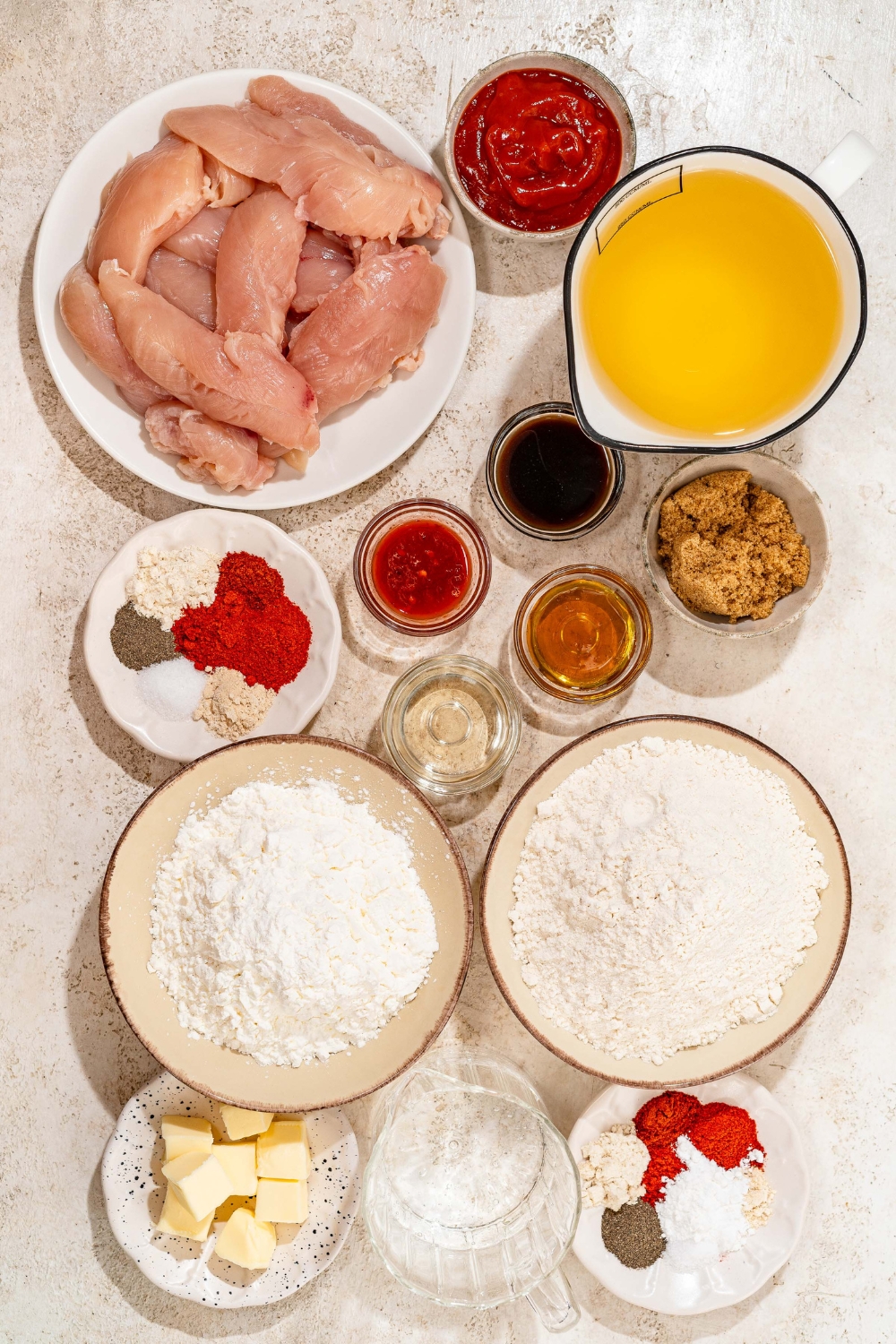 A tan counter with several bowls containing ingredients to make honey BBQ chicken tenders including chicken tenders, oil, flour, cornstarch, brown sugar, apple cider vinegar, hot sauce, honey, butter, and spices.