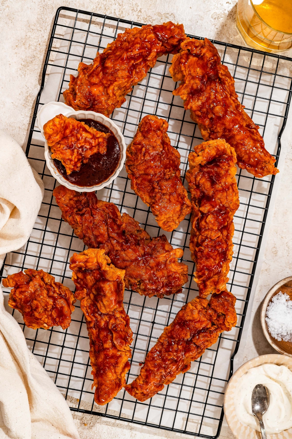 A wire rack with honey BBQ chicken tenders and a bowl of honey BBQ sauce. There is a piece of chicken dipped in the sauce. The rack is on a tan counter.