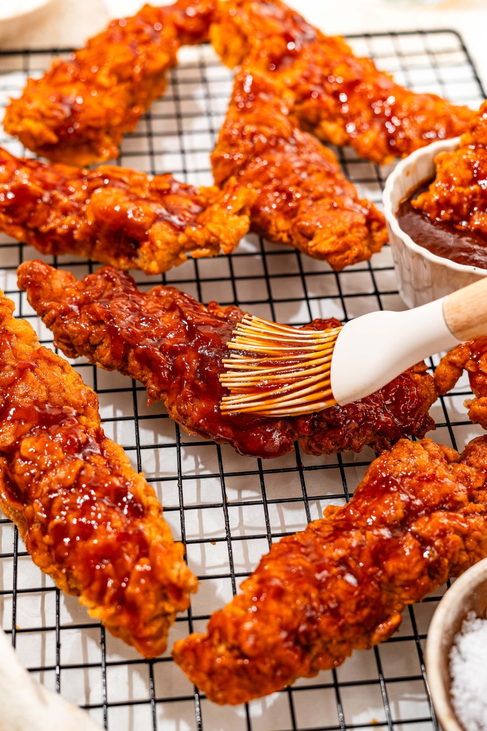 A wire rack with honey BBQ chicken tenders and a bowl of honey BBQ sauce. A brush is brushing the BBQ sauce on one of the tenders.