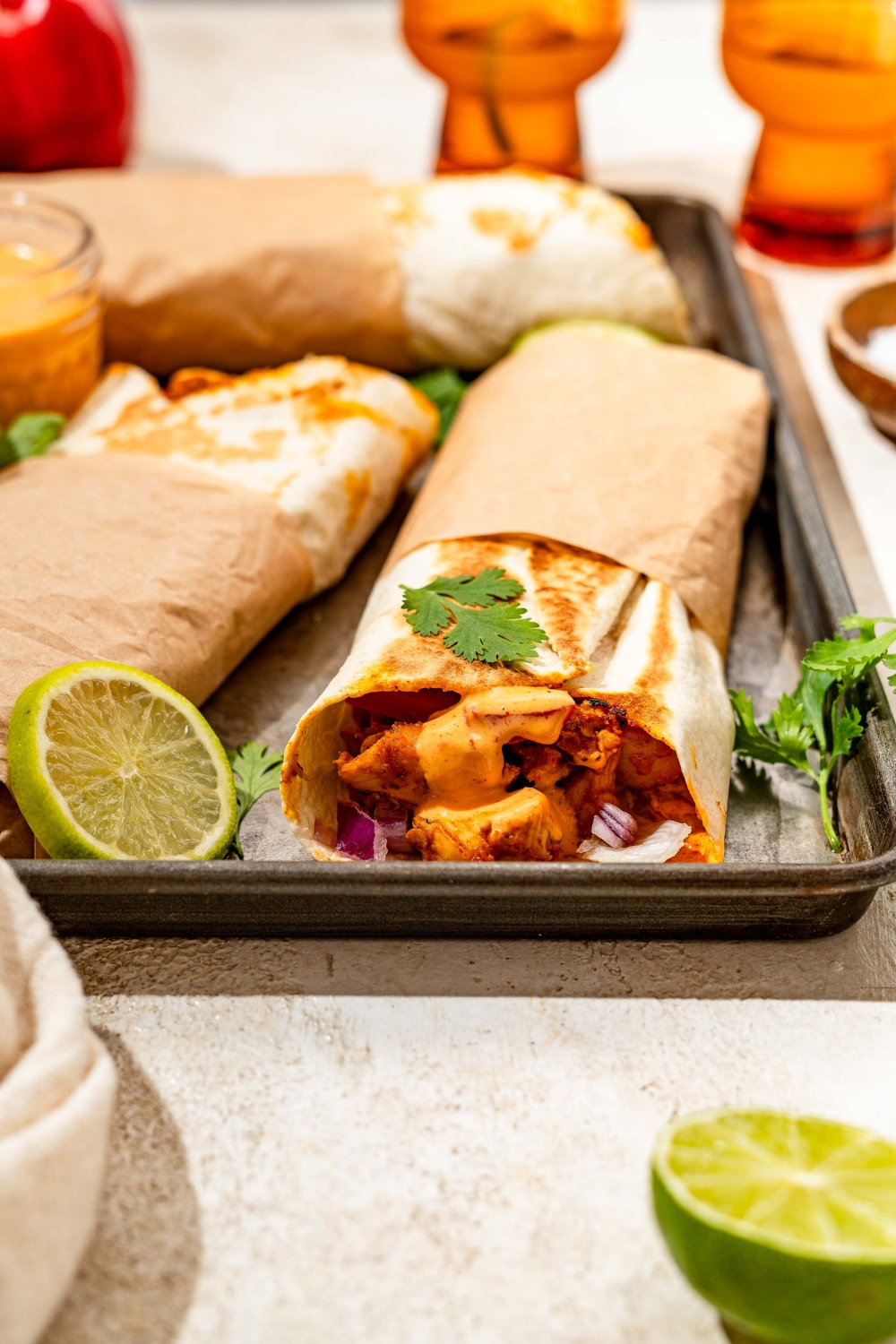 A baking sheet with several honey chipotle chicken wraps wrapped in paper and garnished with cilantro and lime. The sheet is on a tan counter with lime.