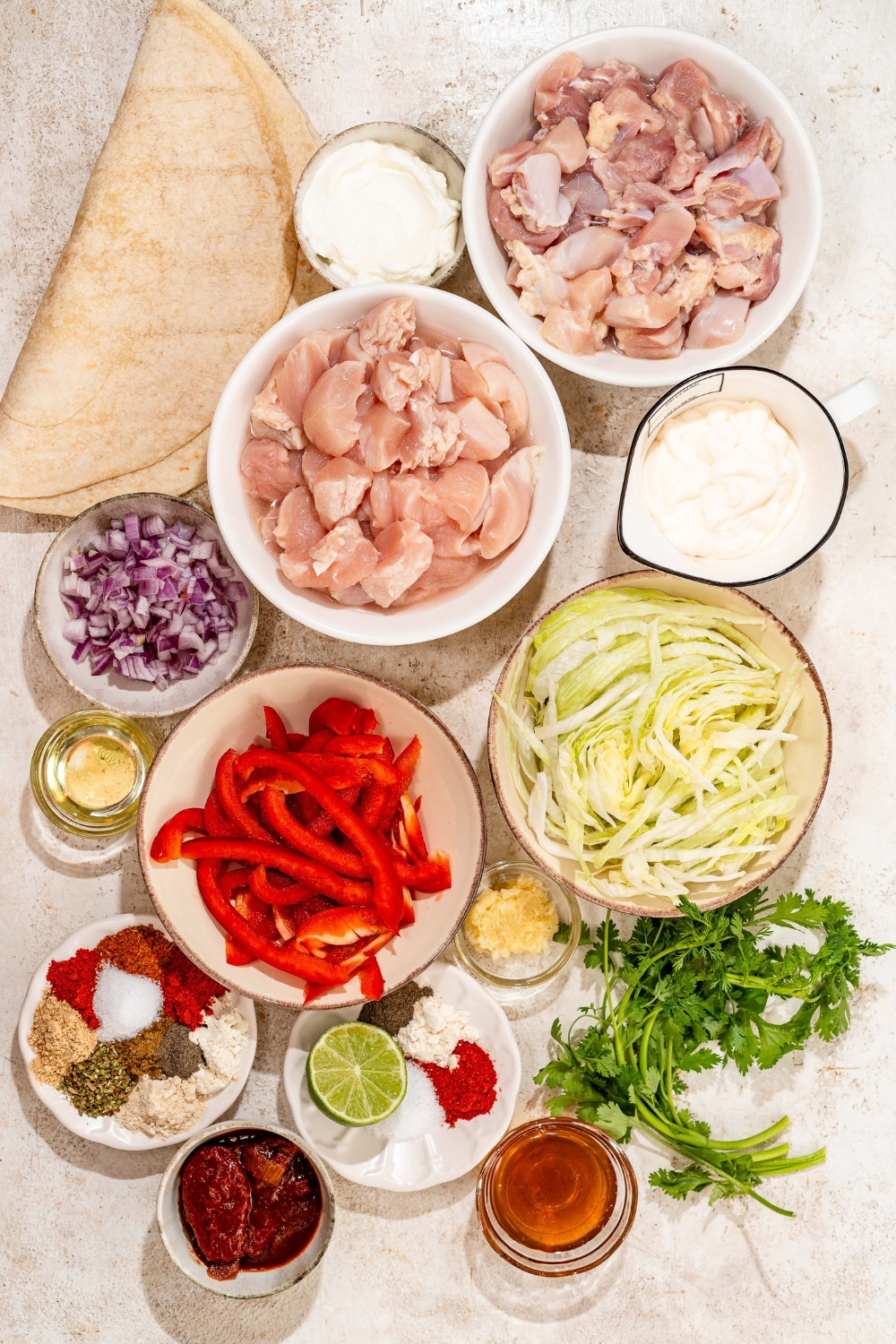 A tan counter with several bowls containing ingredients to make honey chipotle chicken wraps including chicken, tortilla wraps, red pepper, honey, greek yogurt, shredded lettuce, mayo, chipotle adobo sauce, and seasonings.